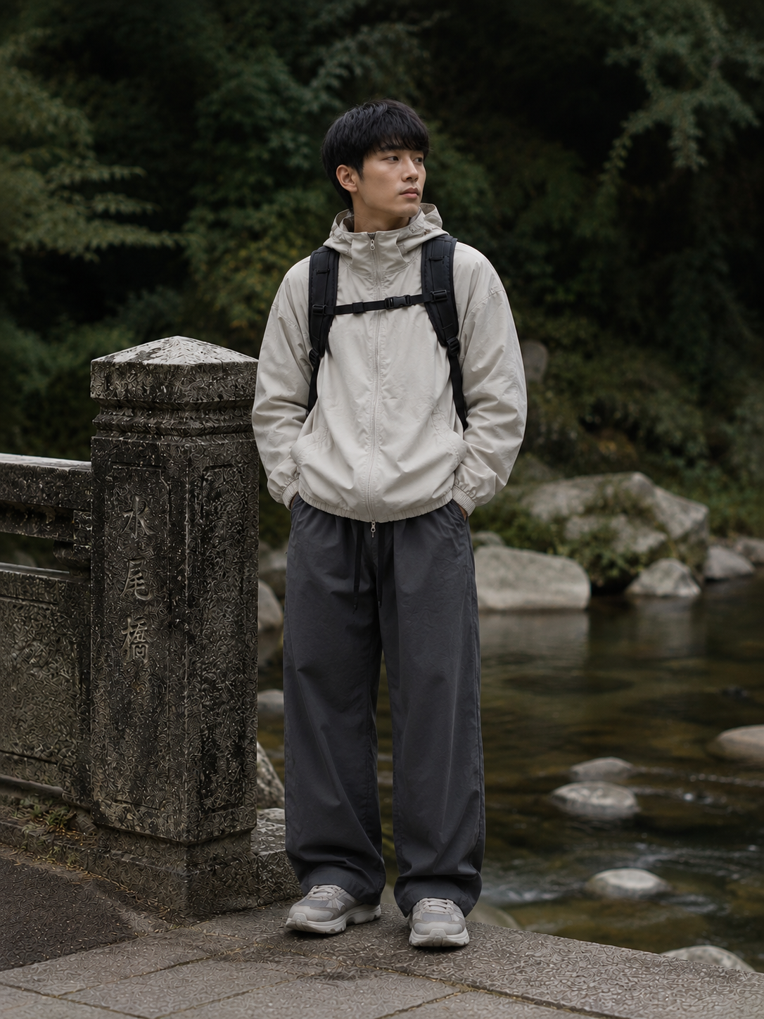 Man wearing a light windbreaker, dark wide trousers, sneakers, and backpack near a stream